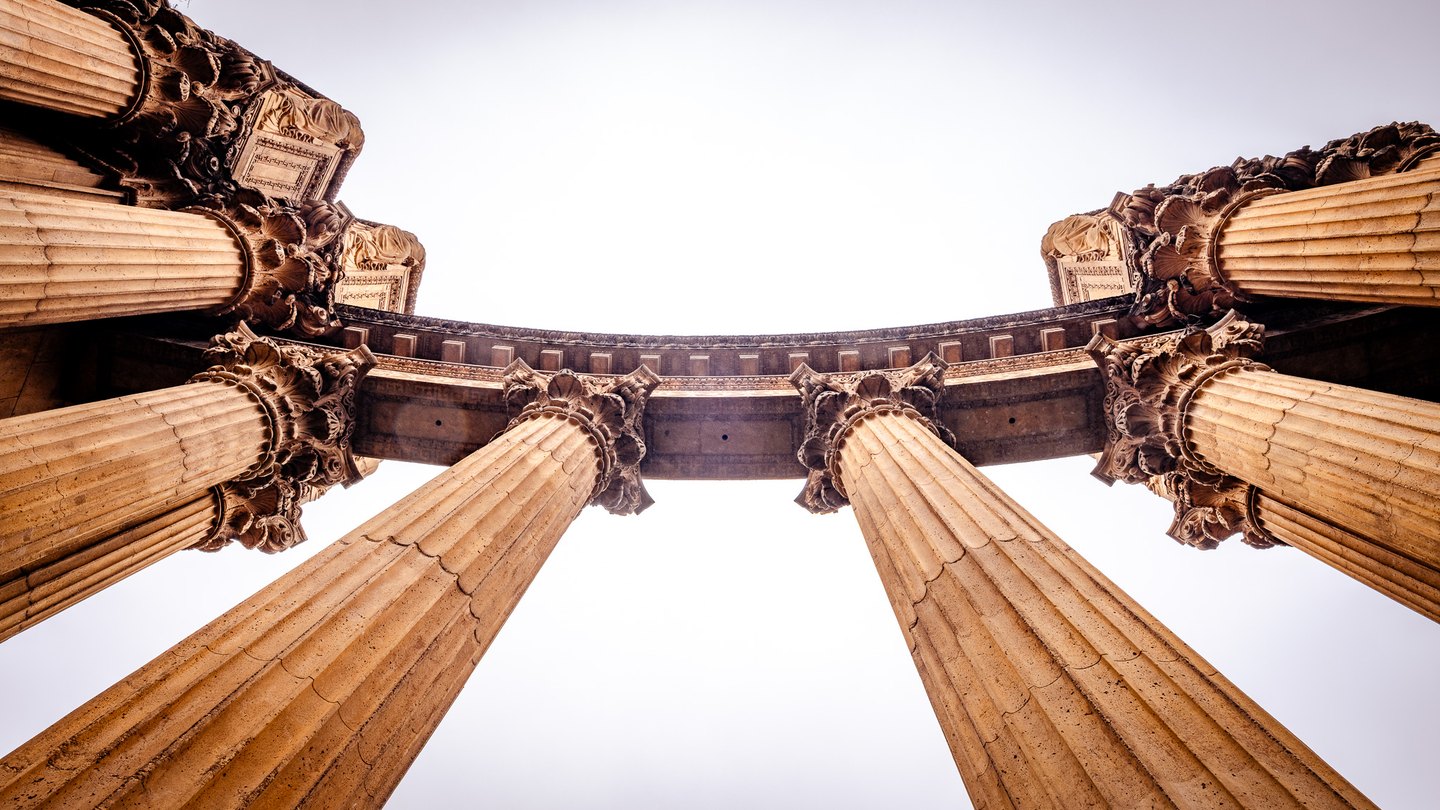 An upward view of fluted columns with ornate capitals supporting a curved entablature against a pale sky.