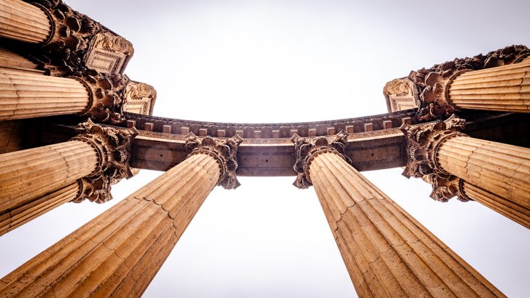 An upward view of fluted columns with ornate capitals supporting a curved entablature against a pale sky.
