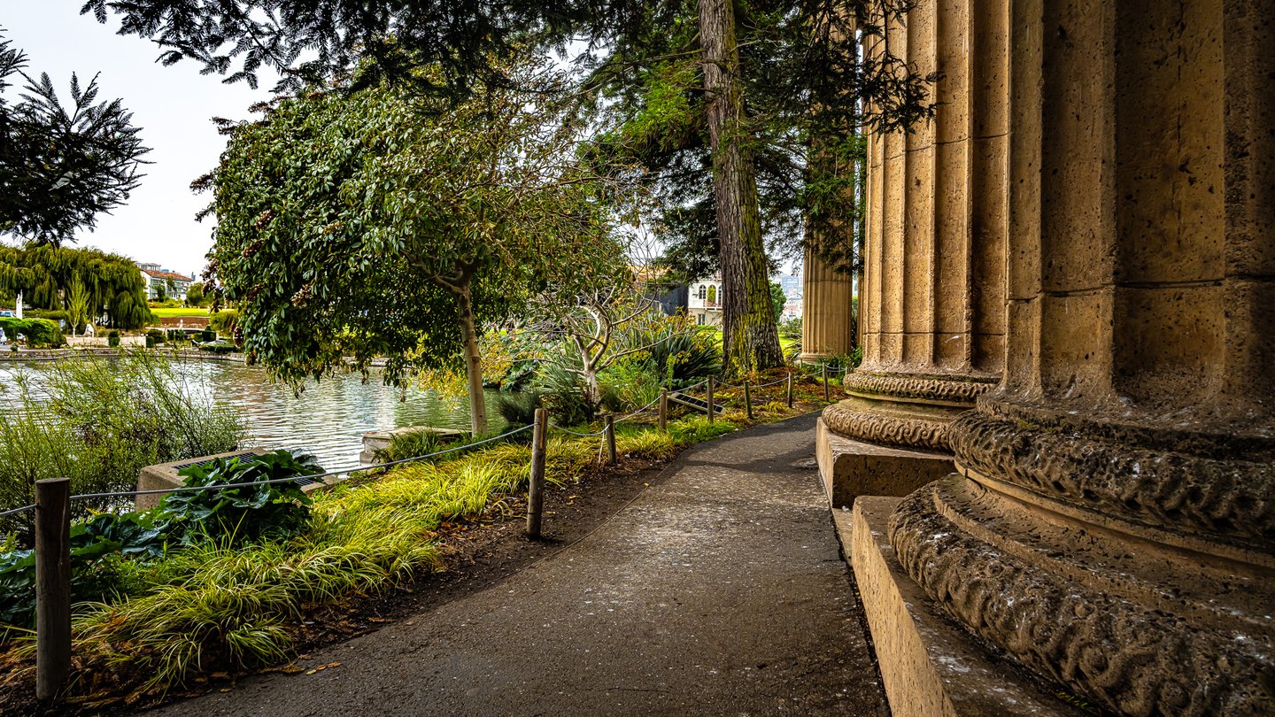 A shaded path curves beside massive fluted columns, with trees, grasses, and a rippling pond to the left.