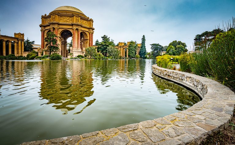 An ornate domed rotunda reflects in a rippling pond bordered by a curved stone wall, tall reeds, and trees.