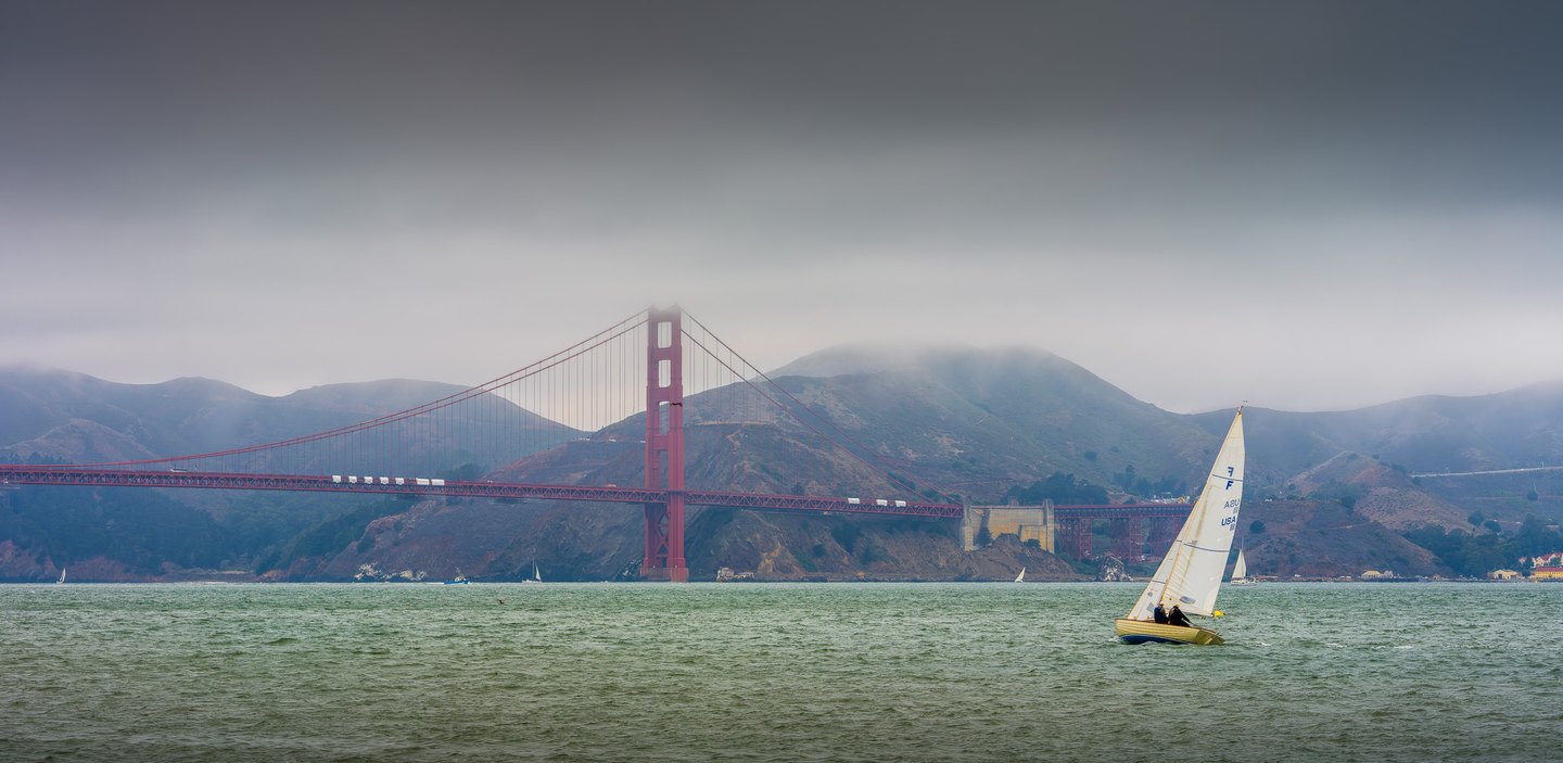 A white-sailed boat crosses rippled green water before a red suspension bridge and hills veiled in low fog.