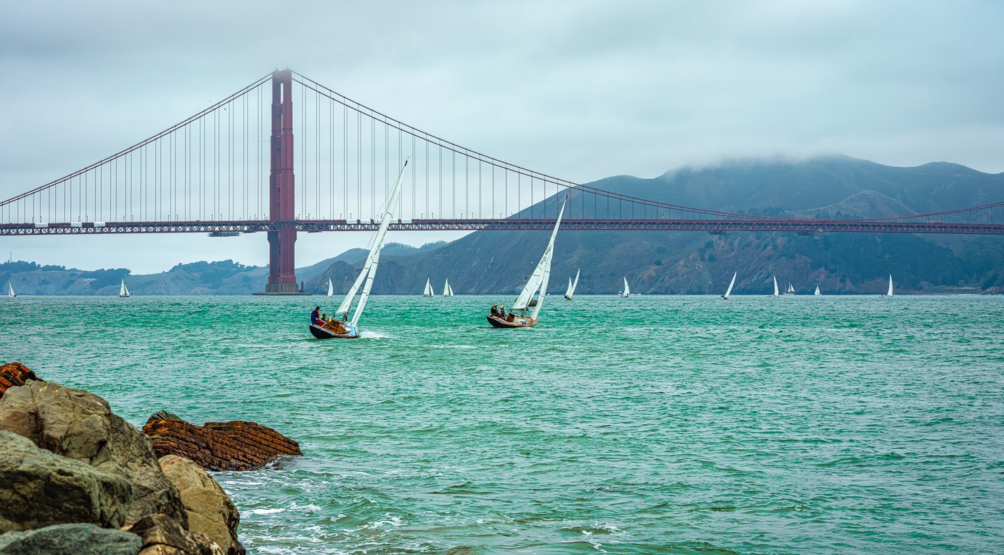 White sailboats cross turquoise water beneath a red suspension bridge, with rocks in front and fog over distant hills.