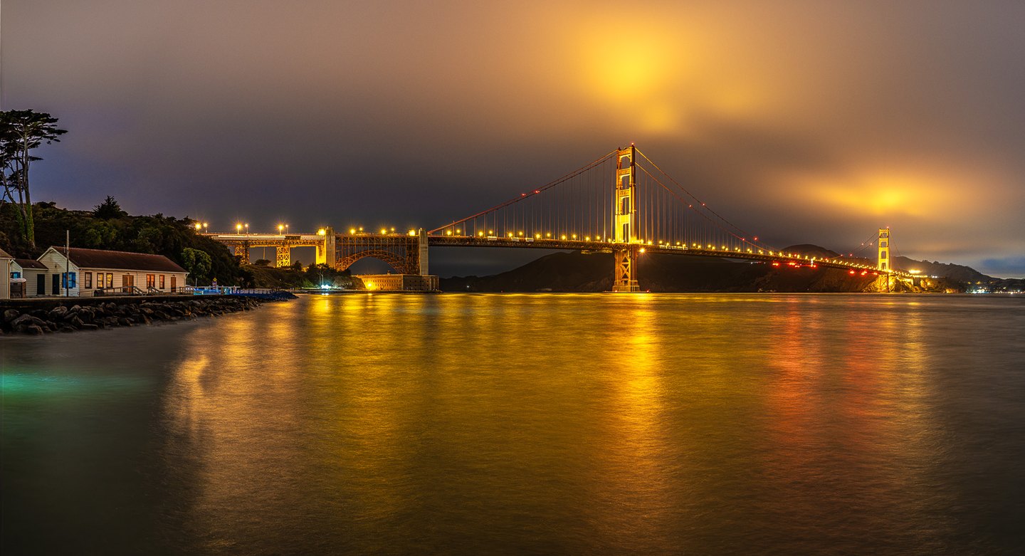 An illuminated suspension bridge spans dark water, casting gold reflections beneath low clouds beside a rocky shore.