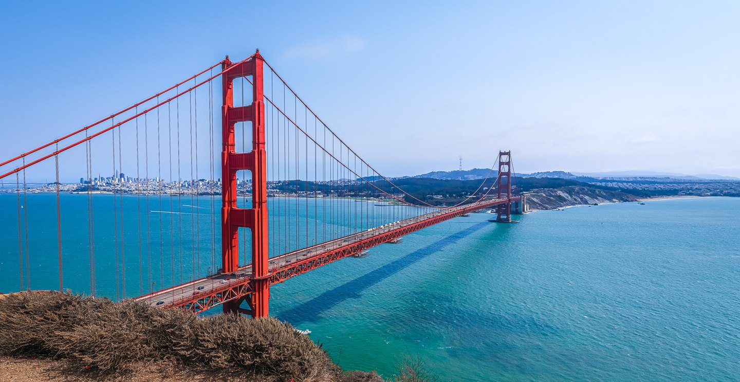 A red suspension bridge crosses turquoise water, with a city skyline beyond and dry shrubs in the foreground.