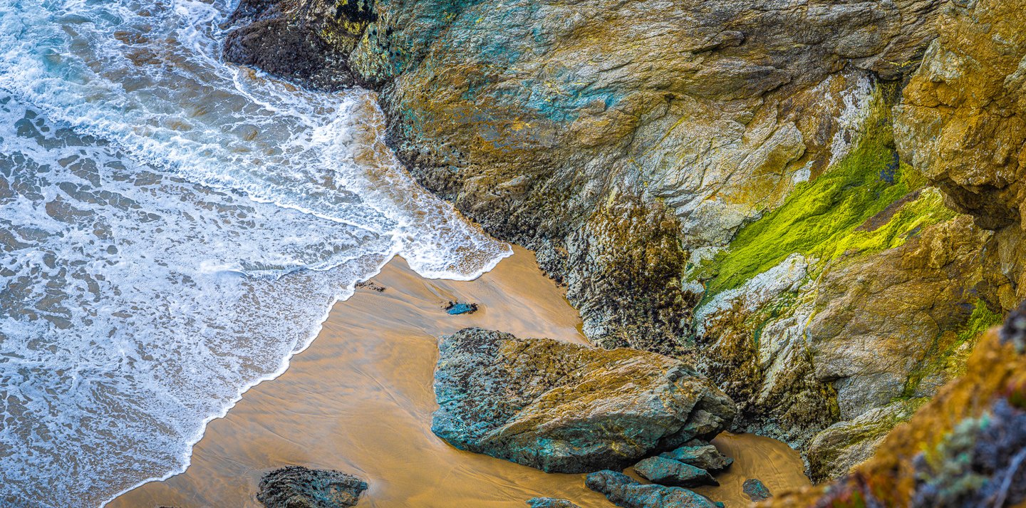 White surf washes golden sand beside rugged coastal rocks streaked with turquoise tones and vivid green growth.