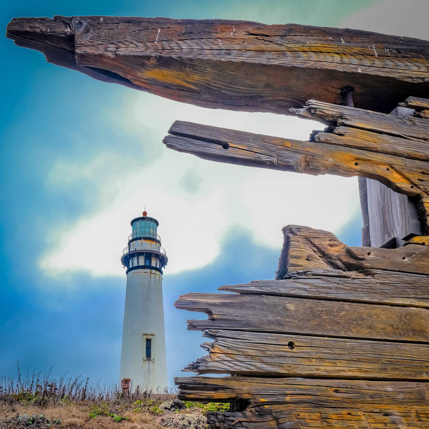 A white lighthouse rises beneath a cloudy blue sky, framed by broken wooden beams and splintered planks.