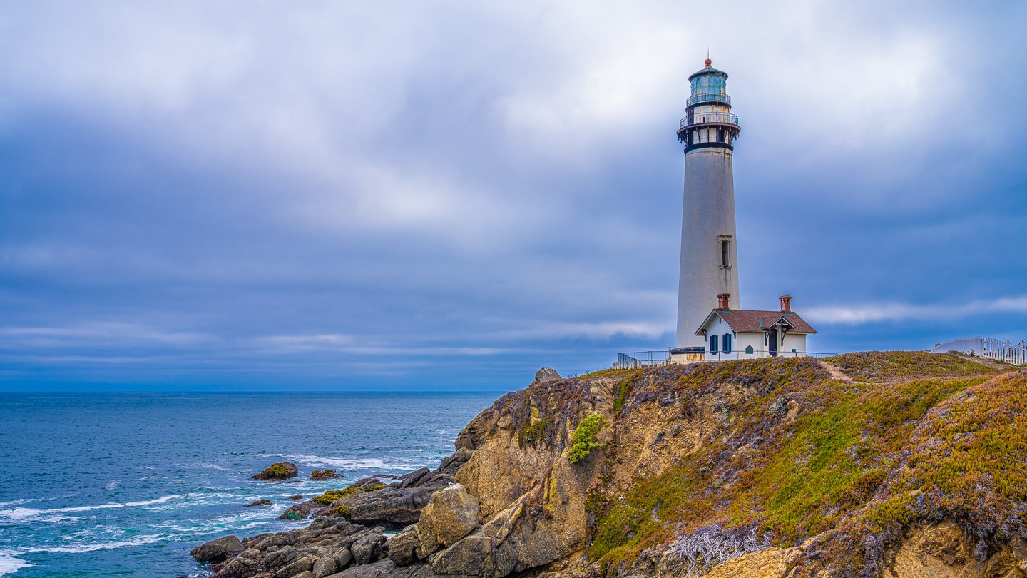 A tall white lighthouse and red-roofed building stand on a rocky coastal bluff beneath blue-gray clouds.