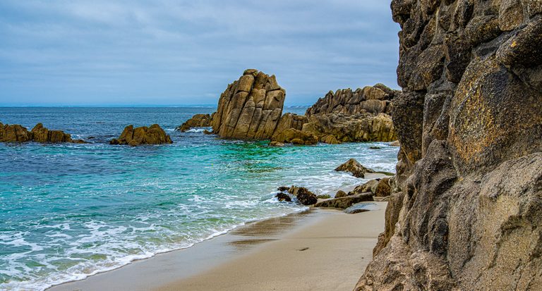 Turquoise waves wash a sandy beach between jagged offshore rocks and a large rock face beneath a cloudy blue sky.