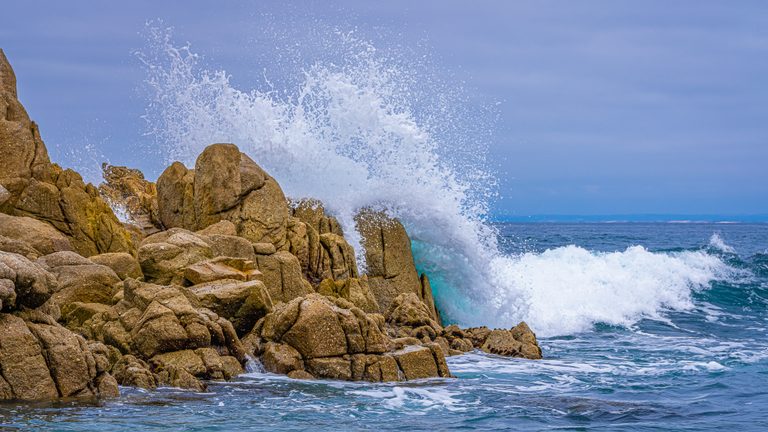 White surf bursts above jagged ochre rocks, with blue sea and a hazy sky beyond.