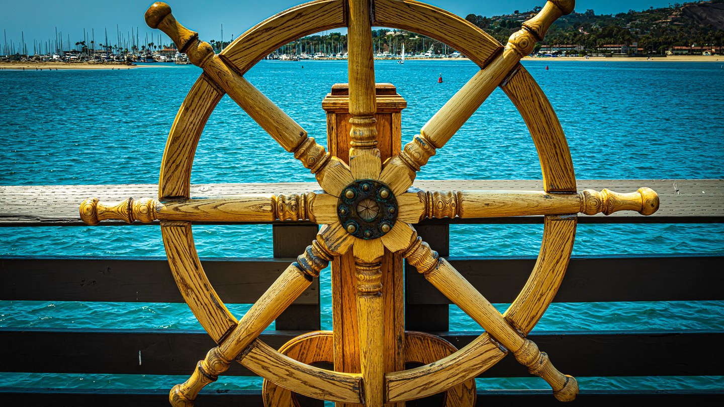 A large wooden ship's wheel stands before turquoise harbor water, with sailboats and a hillside beyond.
