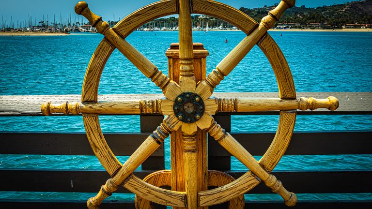 A large wooden ship's wheel stands before turquoise harbor water, with sailboats and a hillside beyond.