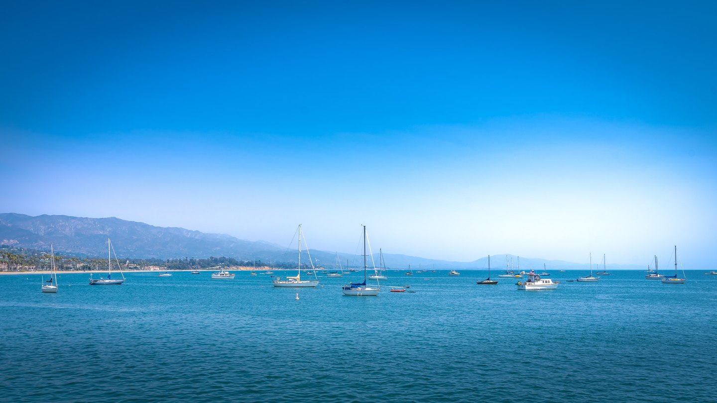 Sailboats and motorboats dot turquoise water beneath a clear blue sky, with mountains and a palm-lined shore beyond.