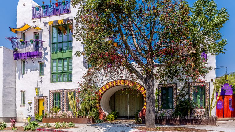 A white building with purple balconies, green windows, and a rounded gateway stands behind a large leafy tree.