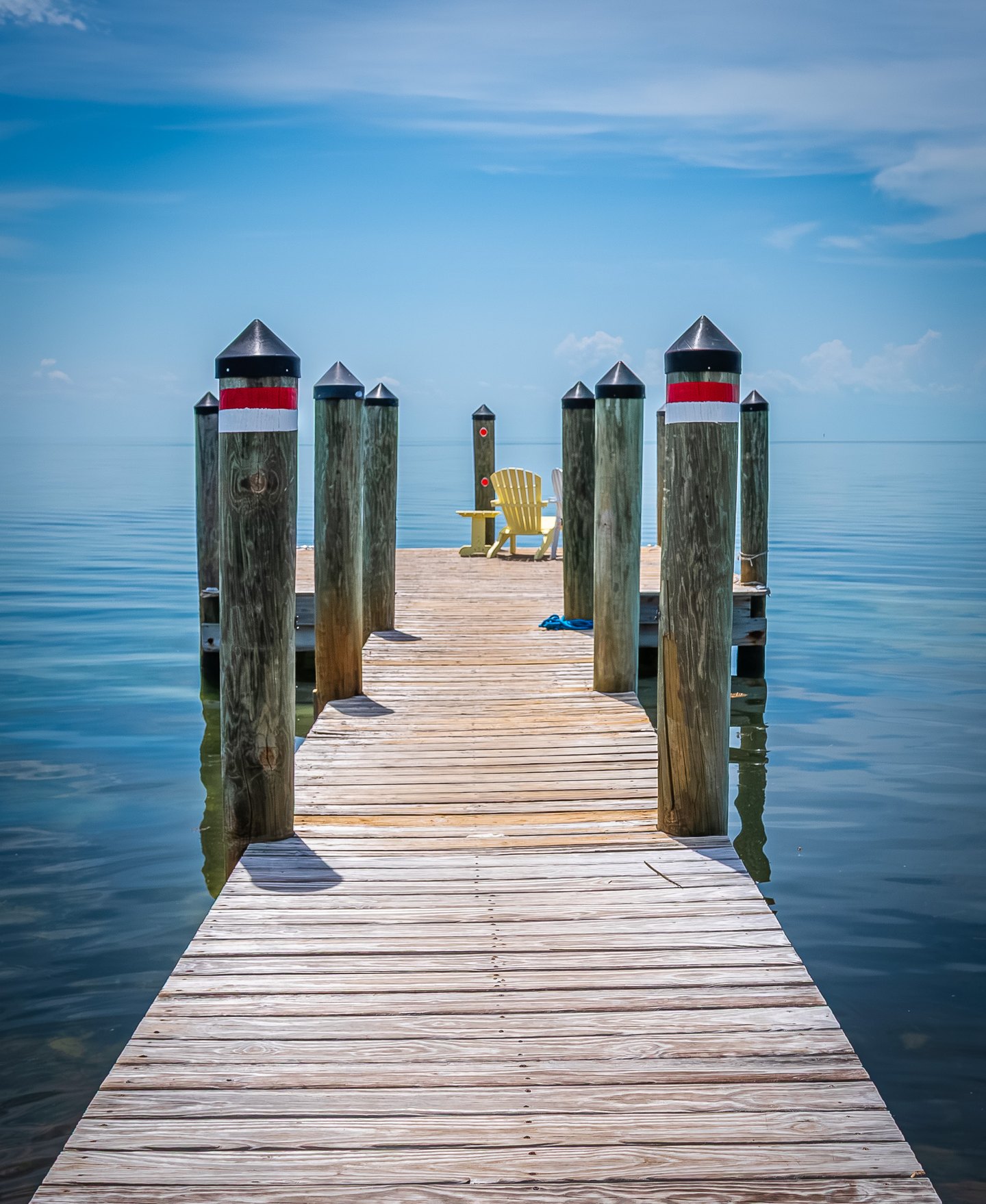 A wooden dock lined with capped pilings leads to a yellow chair above calm blue water beneath a lightly clouded sky.