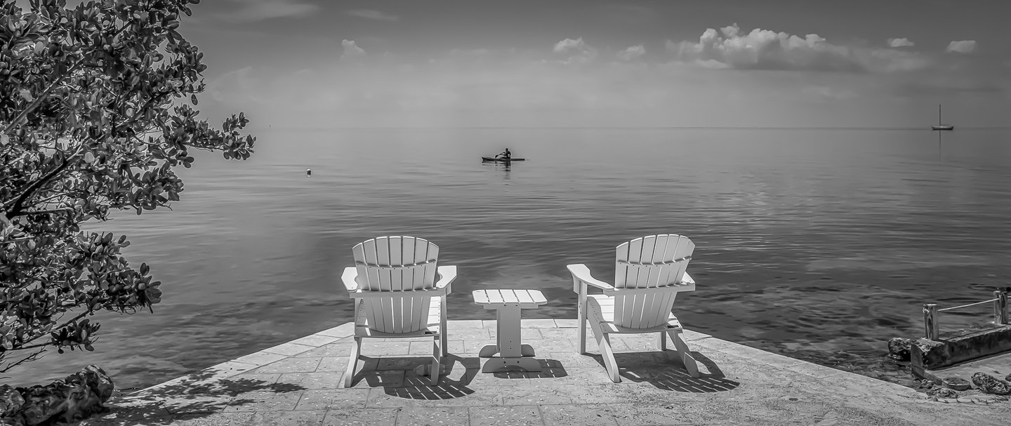 Black-and-white view of two waterfront chairs and a small table, with a paddler and distant sailboat on calm water.