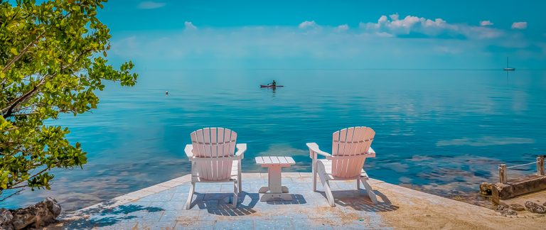 Two pale Adirondack chairs and a small table face calm turquoise water, with a paddler and distant sailboat.