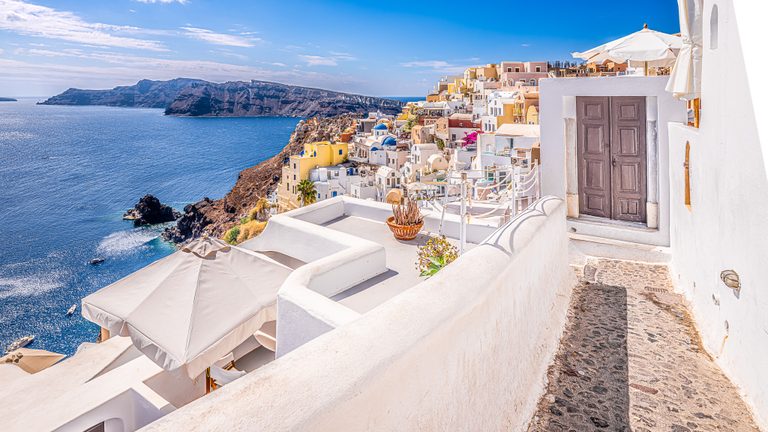 White terraces and a stone path overlook blue water, with pastel cliffside buildings and a rugged ridge beyond.