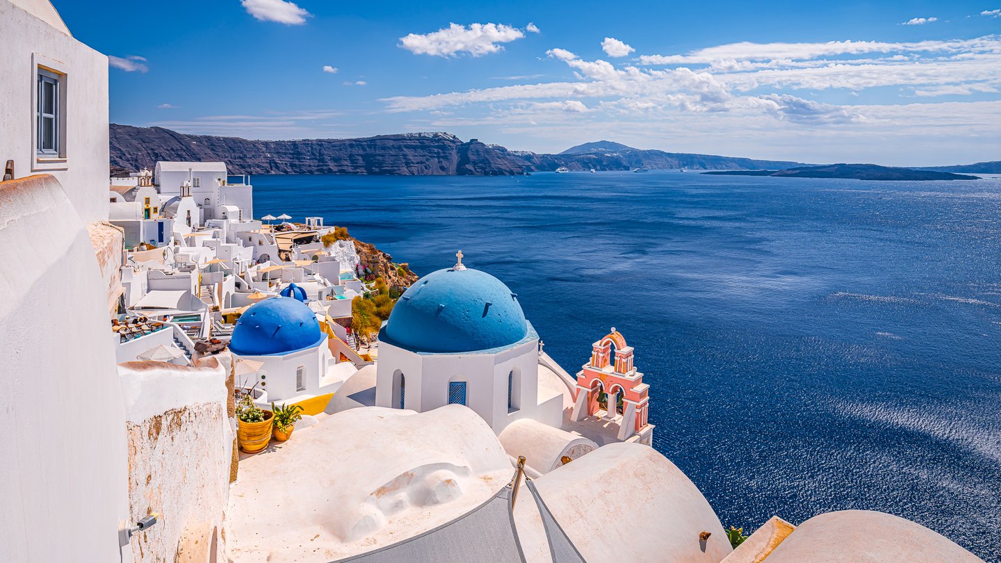 Blue-domed white churches and a pink bell tower overlook deep blue water, distant cliffs, and scattered clouds.