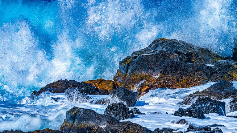Bright blue surf bursts into white spray behind dark coastal rocks, with thick foam swirling through the foreground.