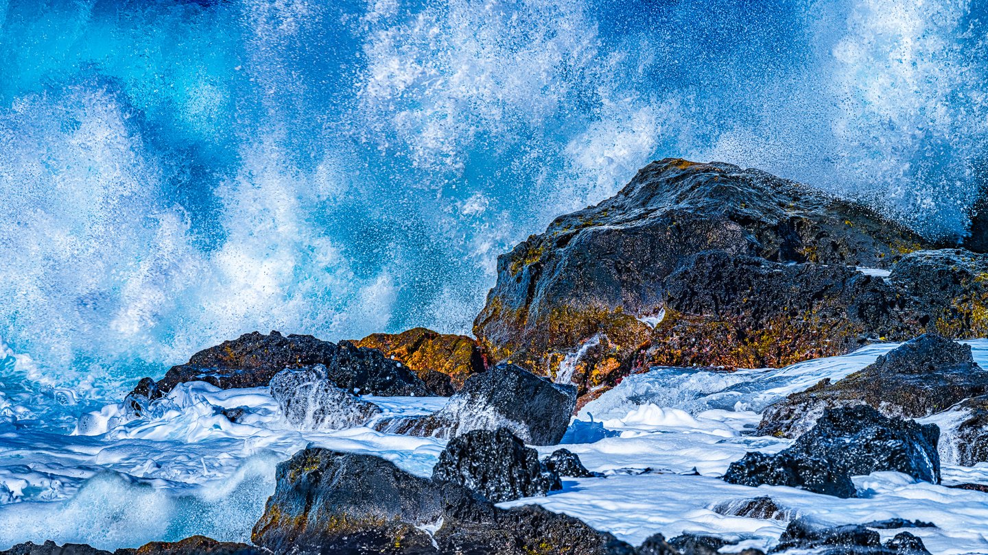 Bright blue surf bursts into white spray behind dark coastal rocks, with thick foam swirling through the foreground.