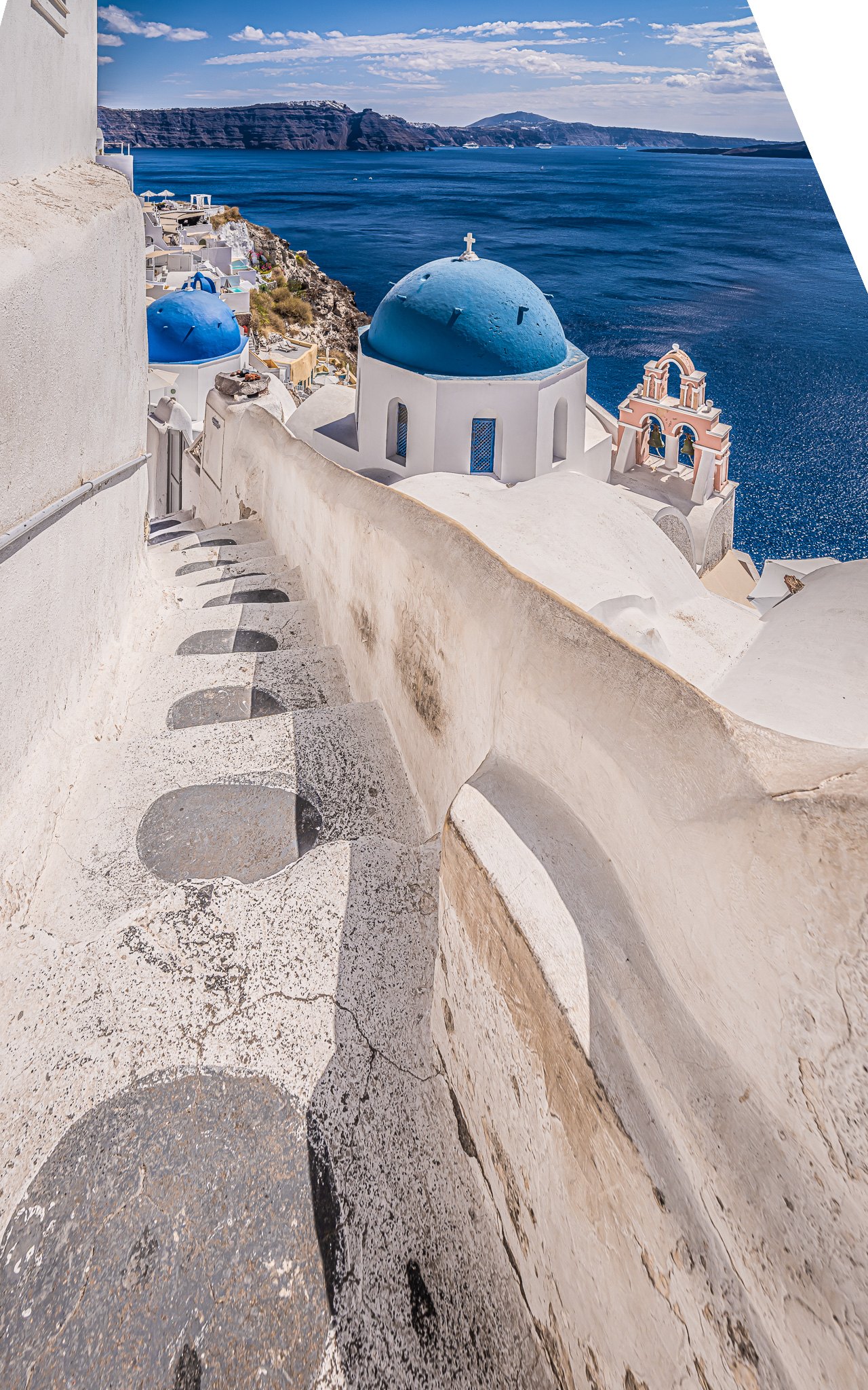 Whitewashed steps descend toward blue-domed churches and a pink bell tower above a deep blue sea and distant cliffs.