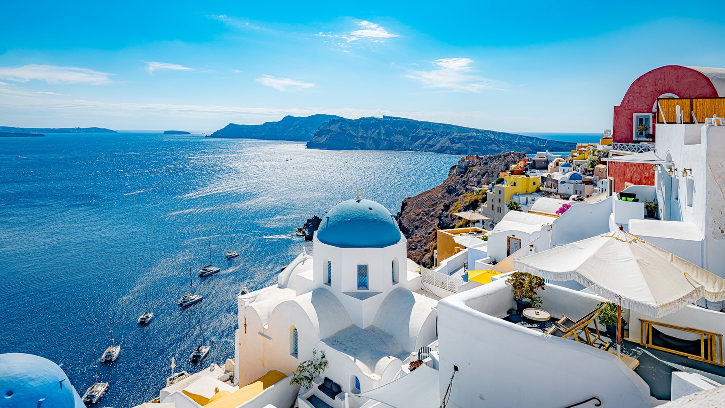 Blue-domed white buildings and sunlit terraces overlook sailboats on a vivid blue sea beneath distant rocky cliffs.