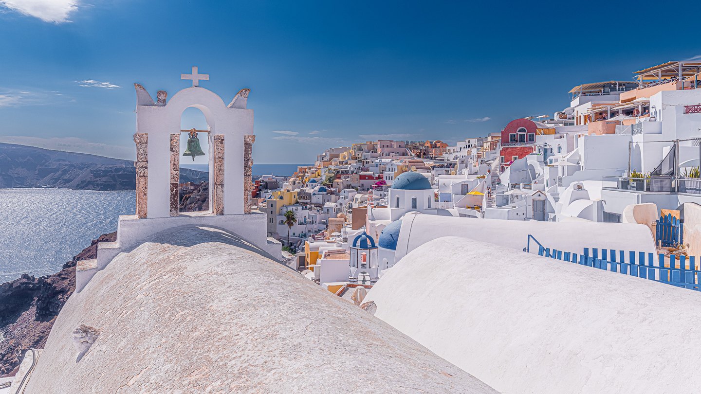 A white bell arch rises above curved rooftops, with blue domes, hillside buildings, and a sunlit sea beyond.