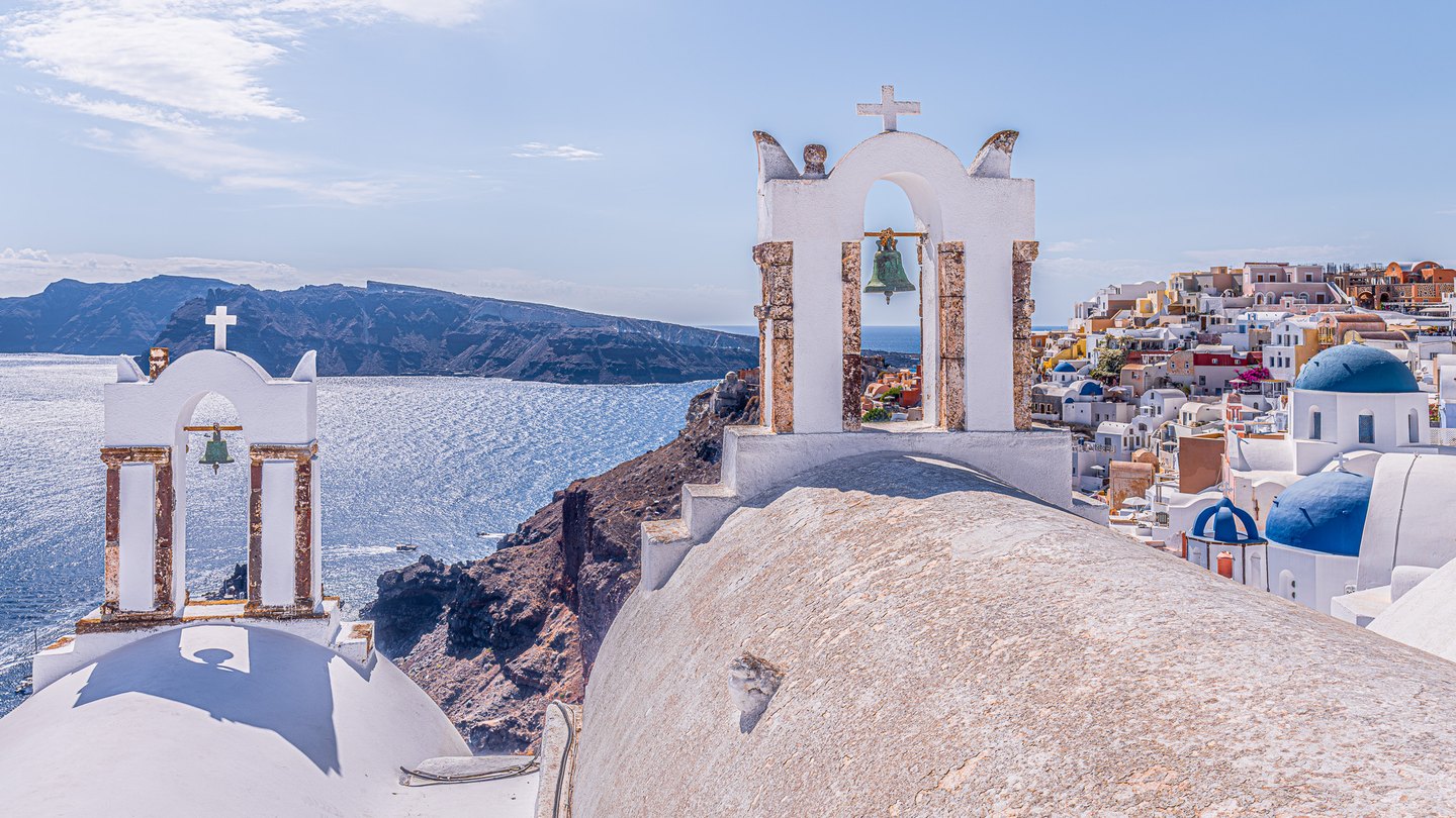 White bell towers and blue-domed churches overlook sparkling sea, rugged cliffs, and a hillside of closely packed buildings.