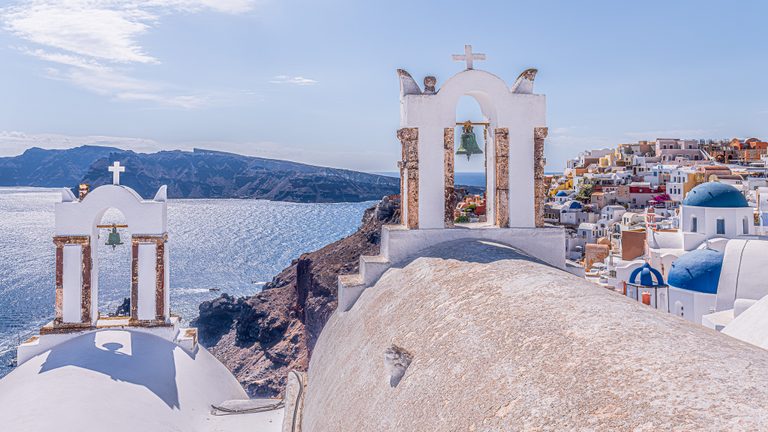 White bell towers and blue-domed churches overlook sparkling sea, rugged cliffs, and a hillside of closely packed buildings.