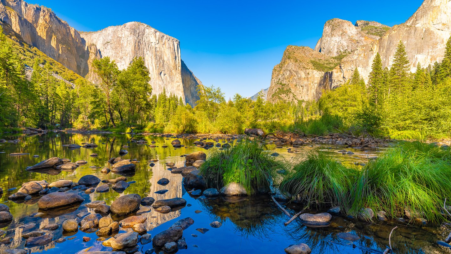 Sunlit cliffs rise above a green forest and a shallow, rocky river reflecting blue sky, trees, and stone.