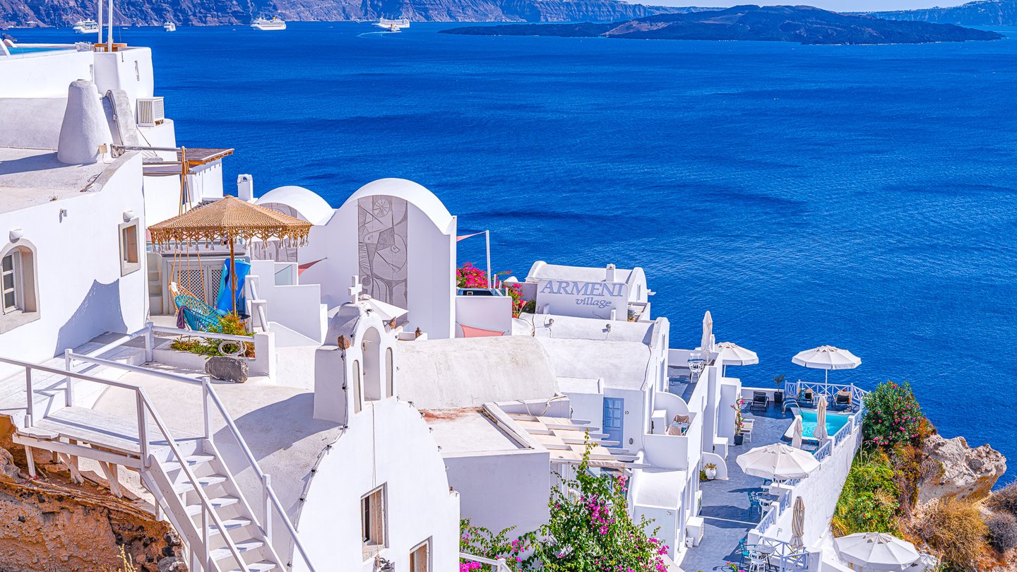 Whitewashed hillside buildings and parasols overlook deep blue water, distant ships, and rugged cliffs.