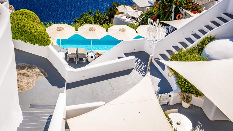 White terraces and stairs surround a turquoise pool with cream umbrellas, green shrubs, and deep blue sea beyond.