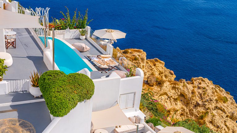 White cliffside terraces with a turquoise pool, loungers, and an umbrella overlook golden rocks and deep blue sea.