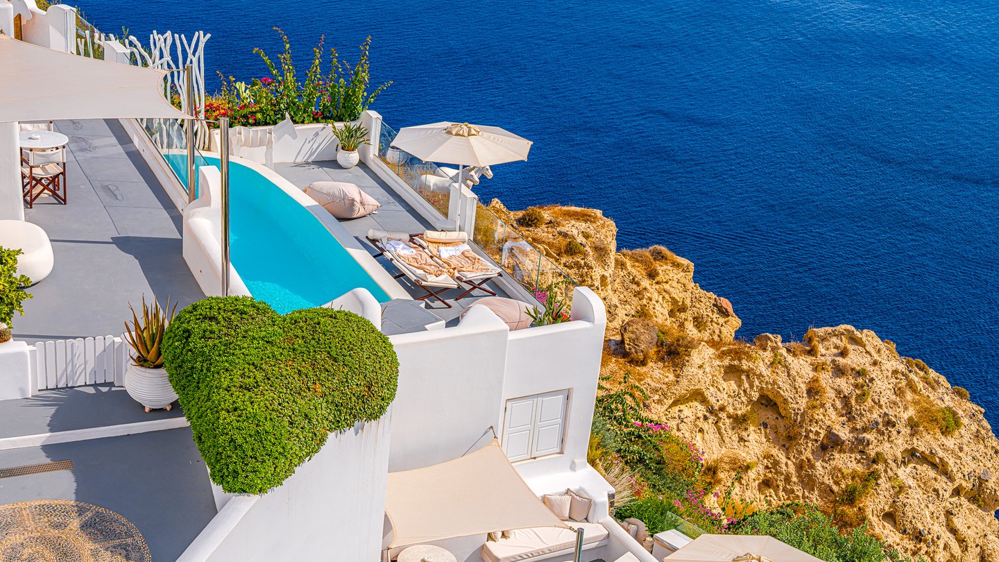 White cliffside terraces with a turquoise pool, loungers, and an umbrella overlook golden rocks and deep blue sea.