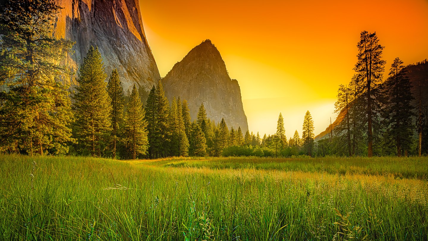 A green meadow and tall conifers lie beneath towering rock cliffs and a vivid orange sky.