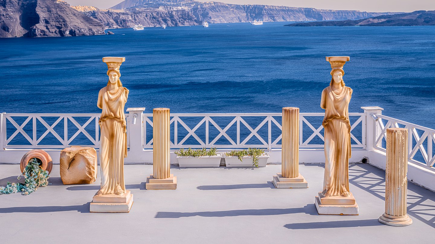 Two draped statues and fluted columns stand on a white-railed terrace above blue water, with ships and cliffs beyond.
