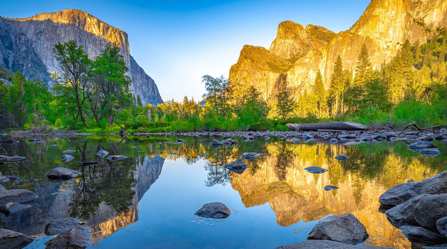 Golden cliffs and green trees reflect in a calm, rock-strewn river beneath a clear blue sky.