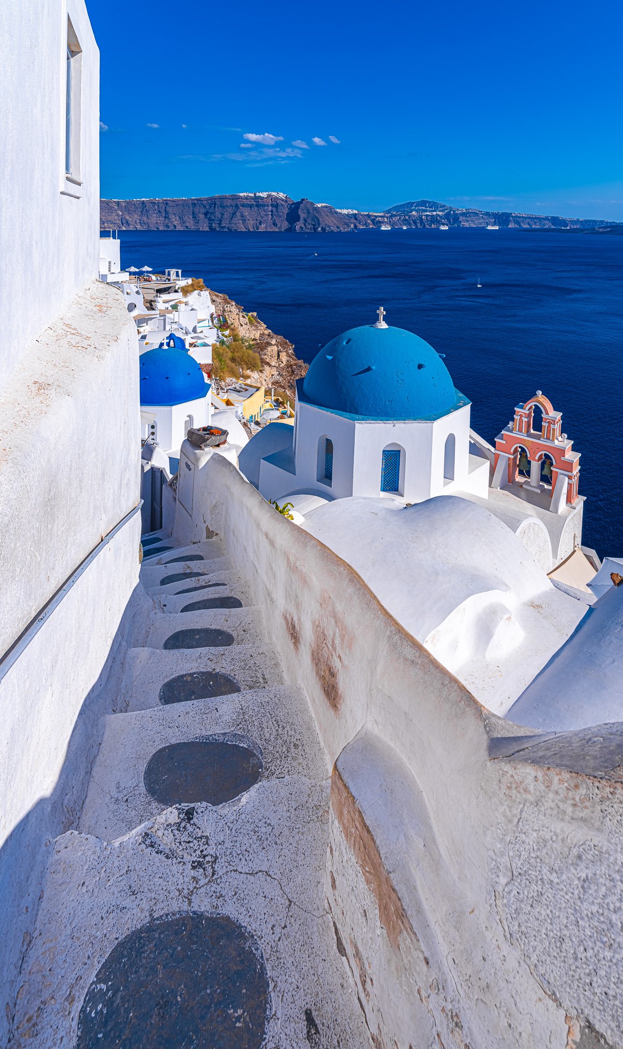 Whitewashed steps descend past blue-domed churches and a coral bell tower toward deep blue sea and distant cliffs.