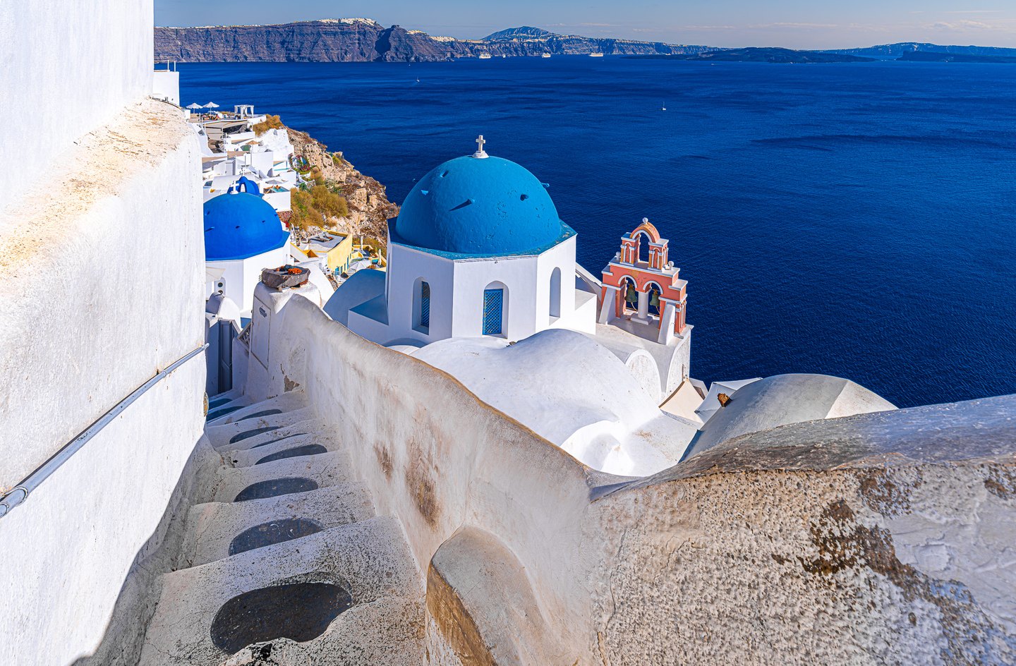 Whitewashed steps descend past blue-domed churches and a peach bell tower above a deep blue sea and distant cliffs.