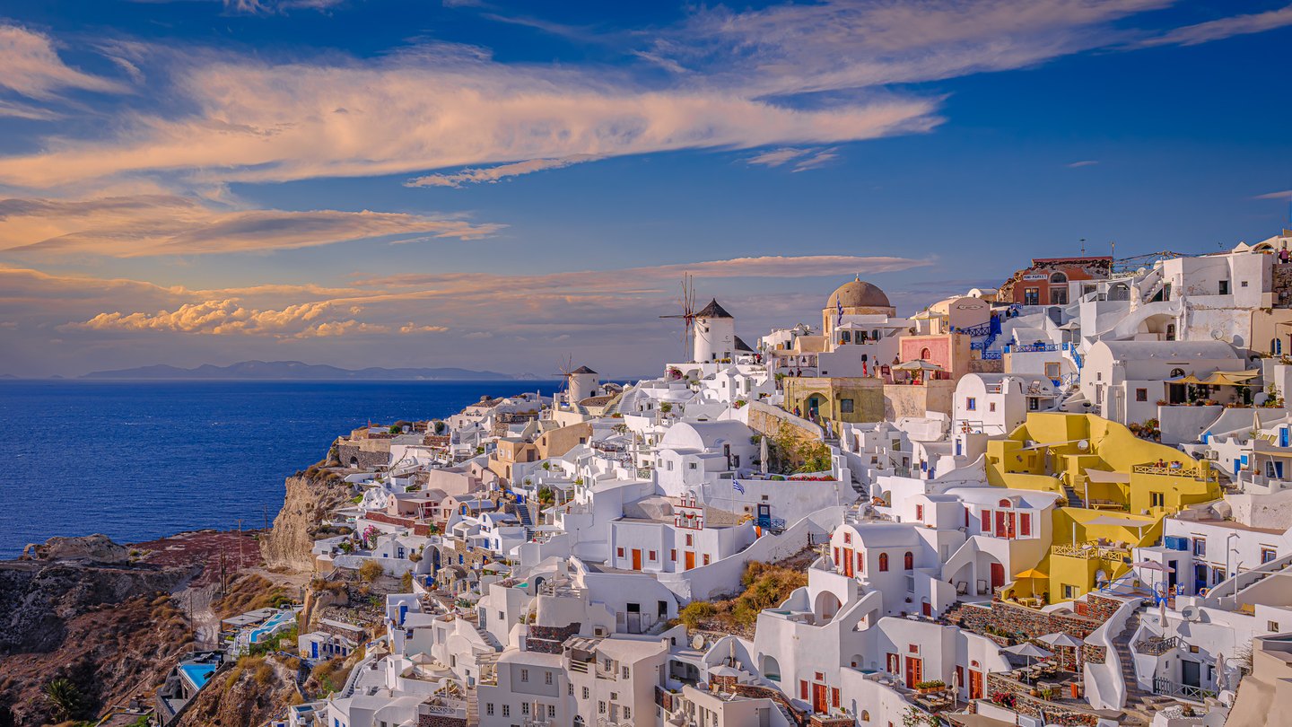 White hillside buildings, two windmills and yellow terraces overlook a deep blue sea beneath peach-tinted clouds.