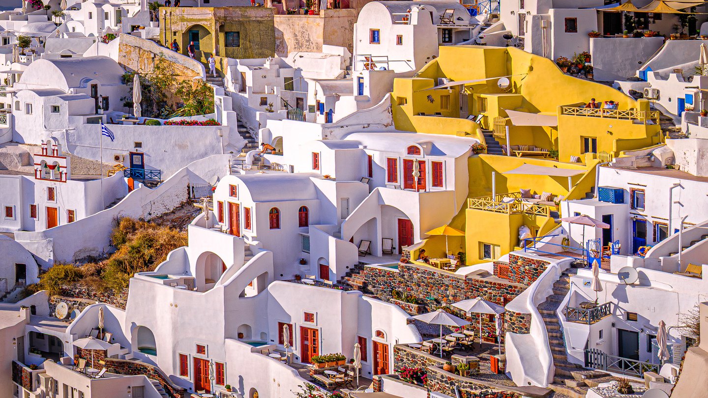 White and yellow hillside buildings overlap with winding stairs, terraces, colorful doors, umbrellas, and small figures.
