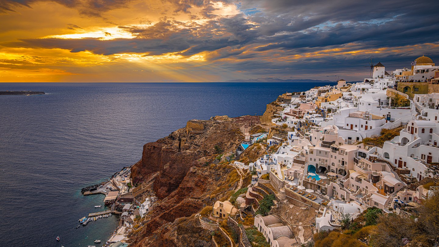 White buildings and windmills crowd a steep coastal cliff above blue water beneath gold and slate sunset clouds.