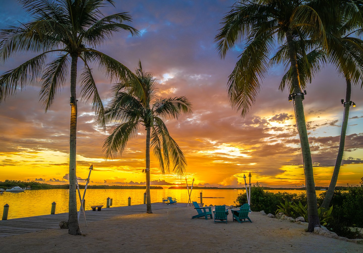 Palm trees frame a golden sunset over calm water, with a wooden dock and turquoise chairs on the sandy shore.