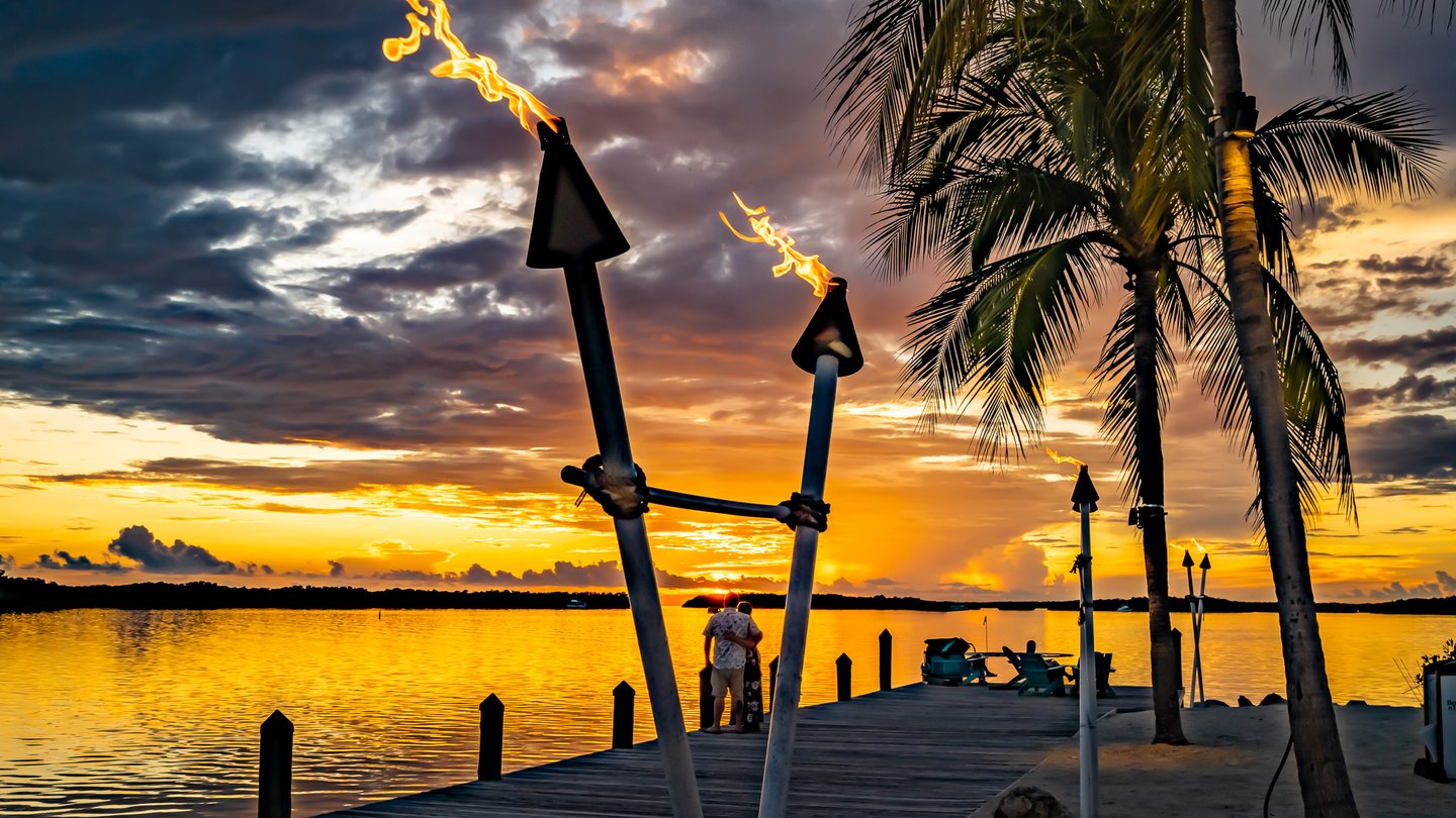 Flaming torches and palms frame two people on a wooden dock beside golden water beneath a clouded sunset sky.