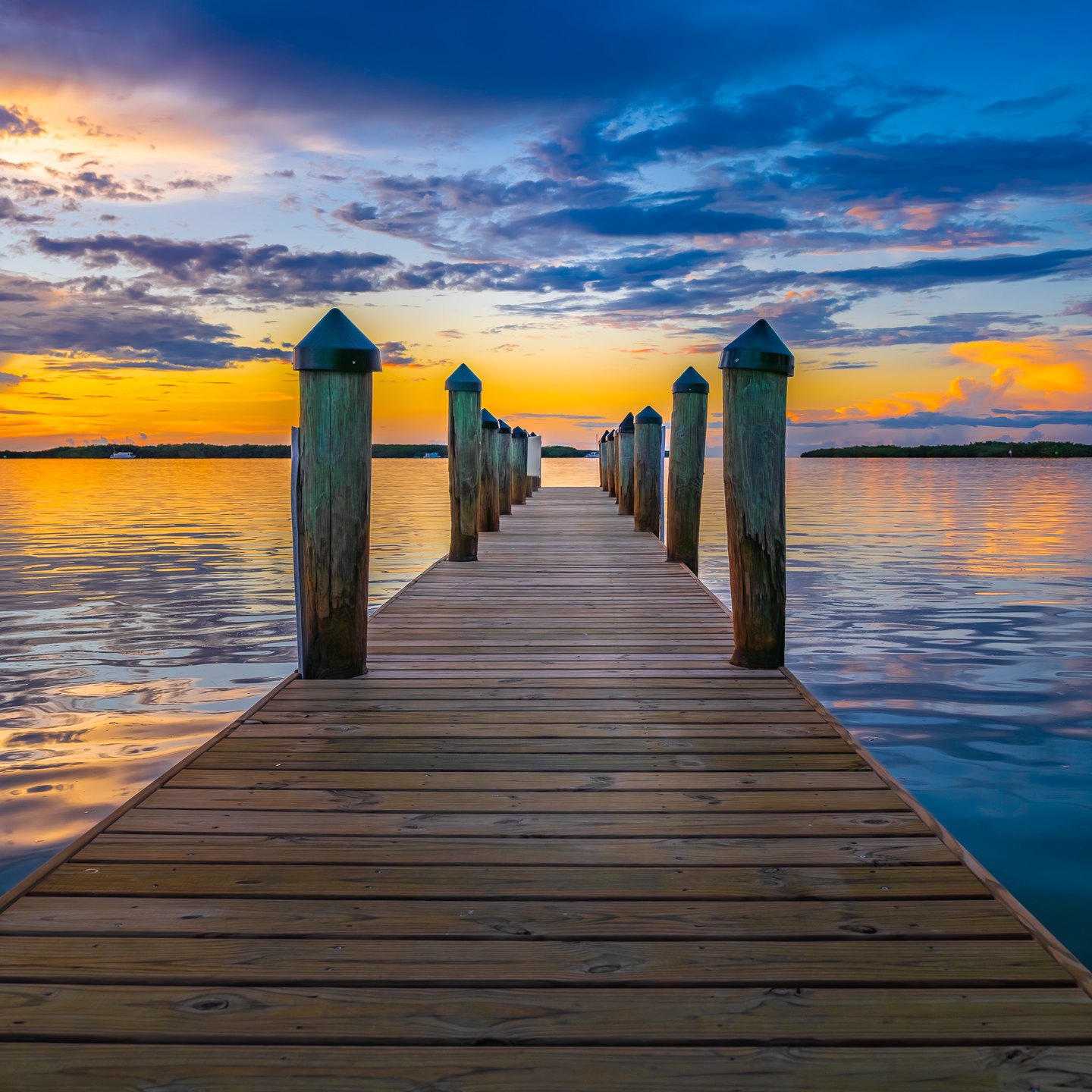 A wooden pier with capped posts extends into calm water beneath a gold and blue, cloud-filled sky.
