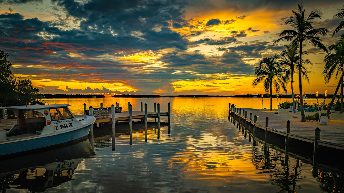 A moored boat and palm-lined docks frame calm water reflecting a golden sunset beneath dark clouds.