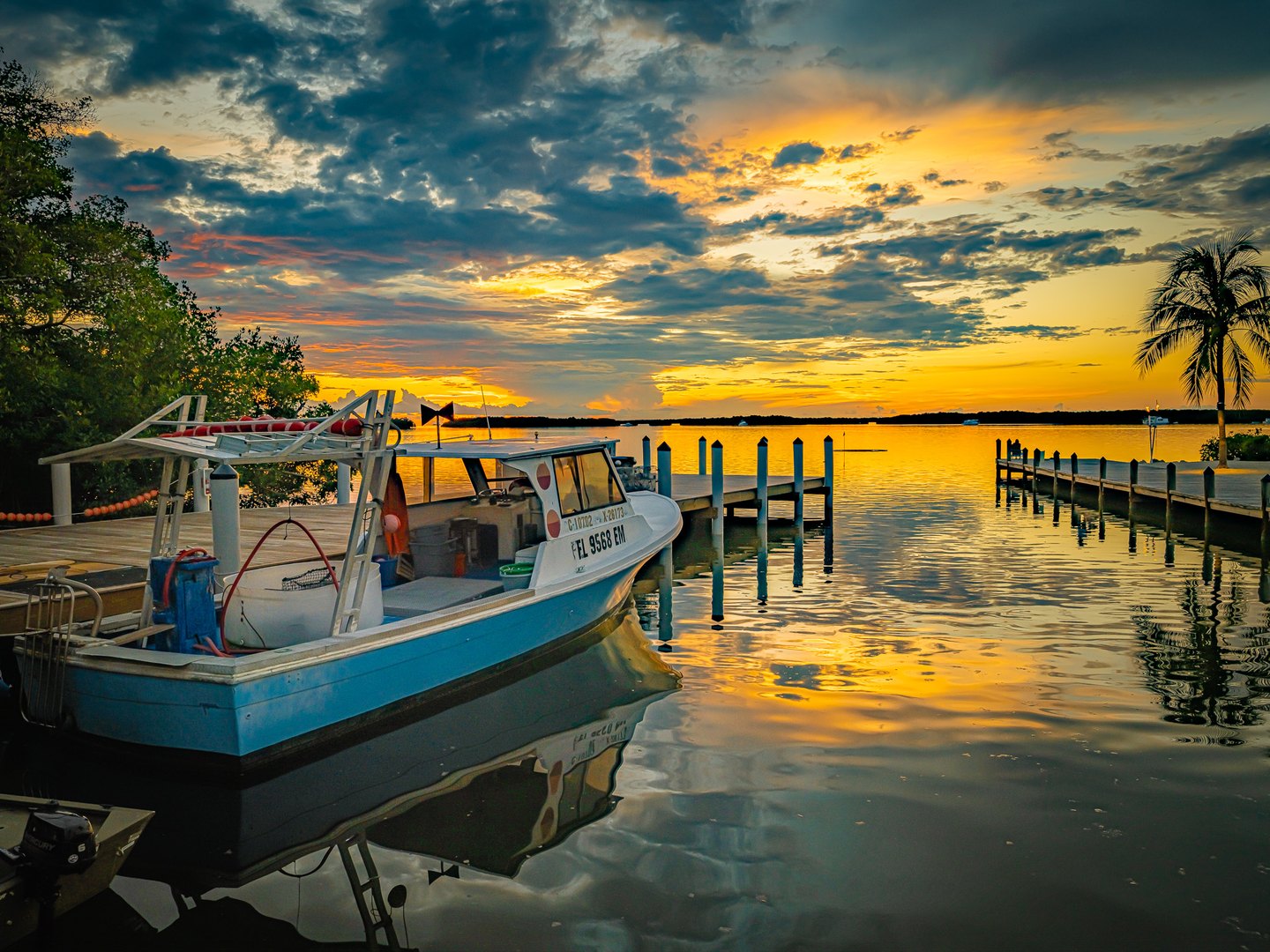 A blue-and-white boat rests beside wooden docks beneath a golden, cloud-filled sky, with a palm on the right.