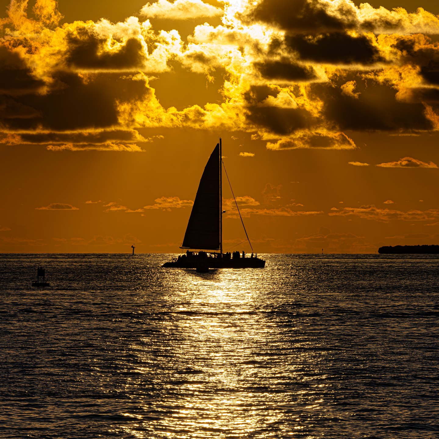 A silhouetted sailboat with passengers crosses golden water beneath dark clouds edged in brilliant sunlight.