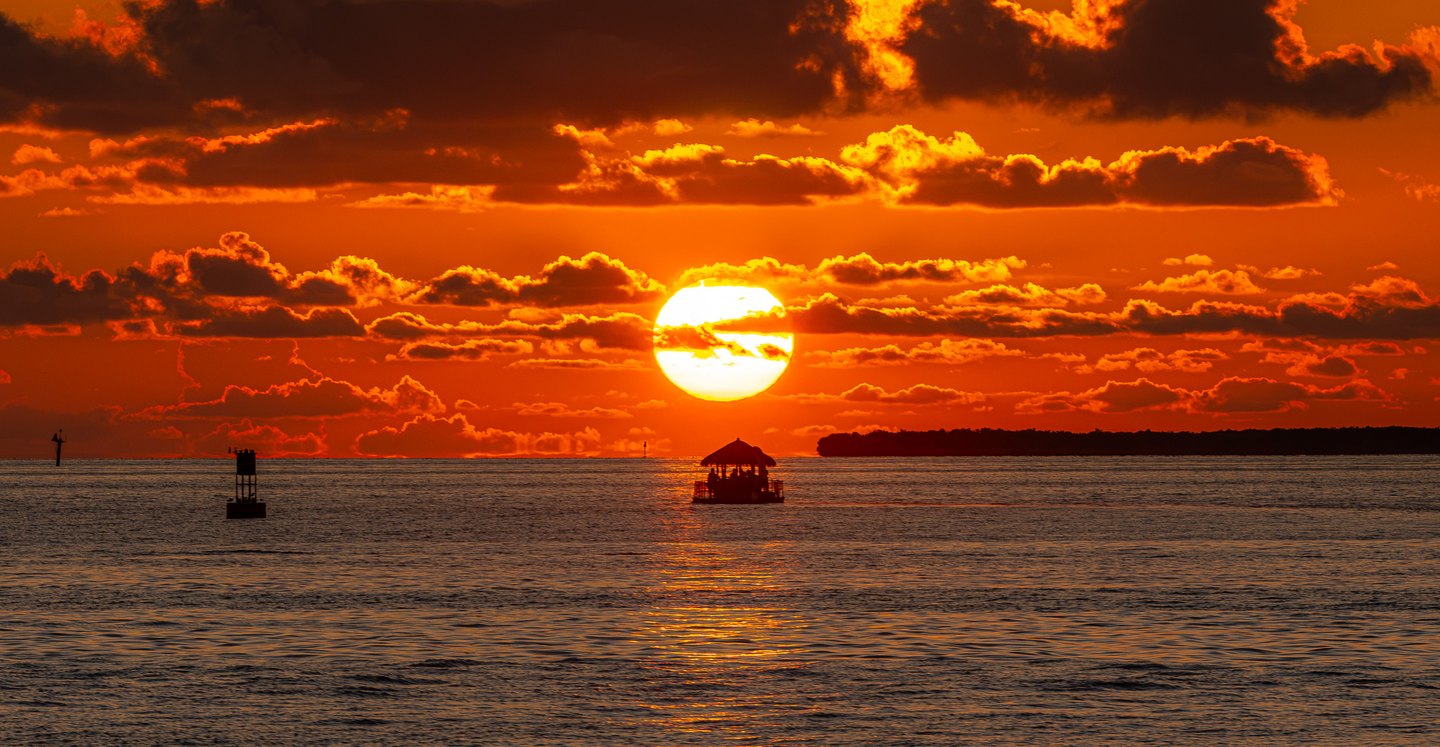 An orange sunset illuminates clouds and rippled water above a silhouetted thatched-roof boat and channel markers.