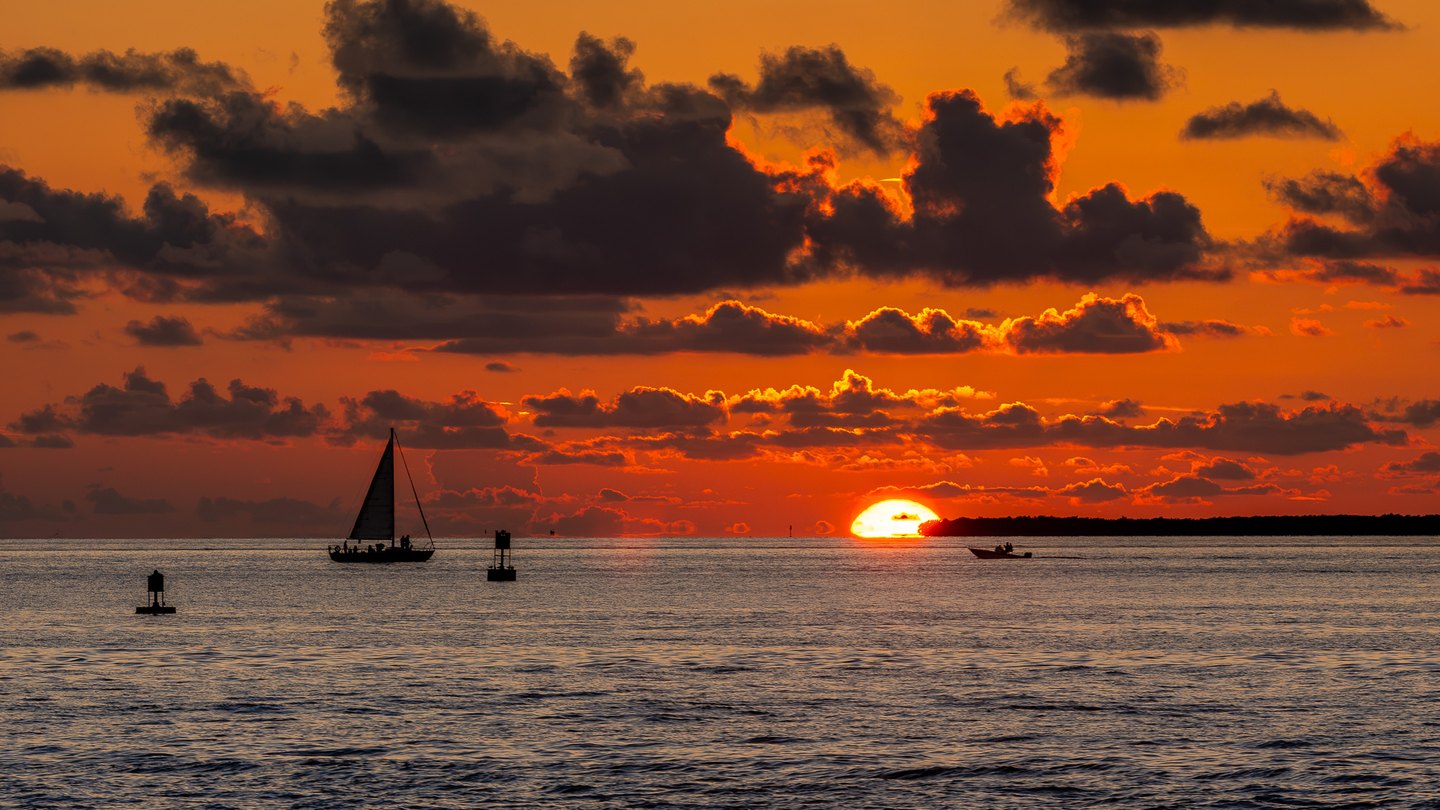 A sailboat and motorboat silhouette against an orange sunset, with dark clouds, channel markers, and rippled water.